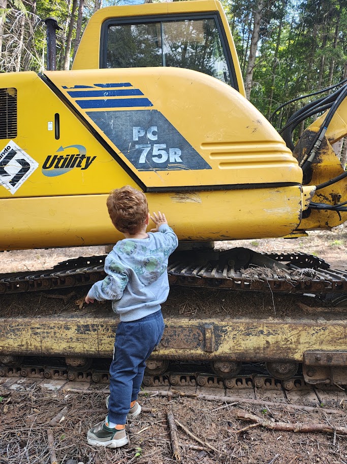 Un bambino tocca con la mano il cingolo di un grande escavatore giallo nel bosco