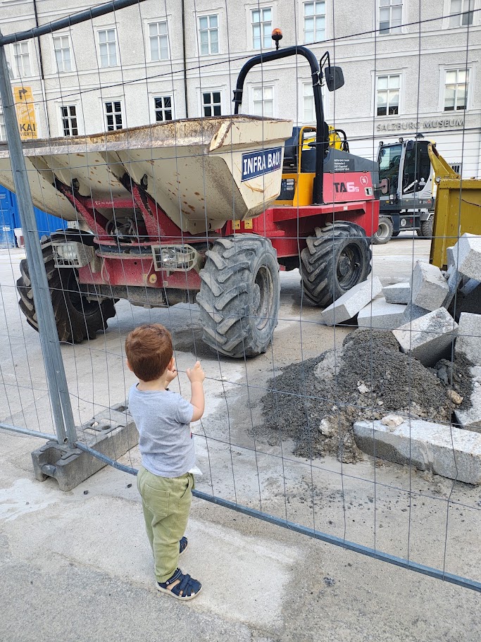 Un bambino indica un dumper e una pala meccanica attraverso la rete di un cantiere