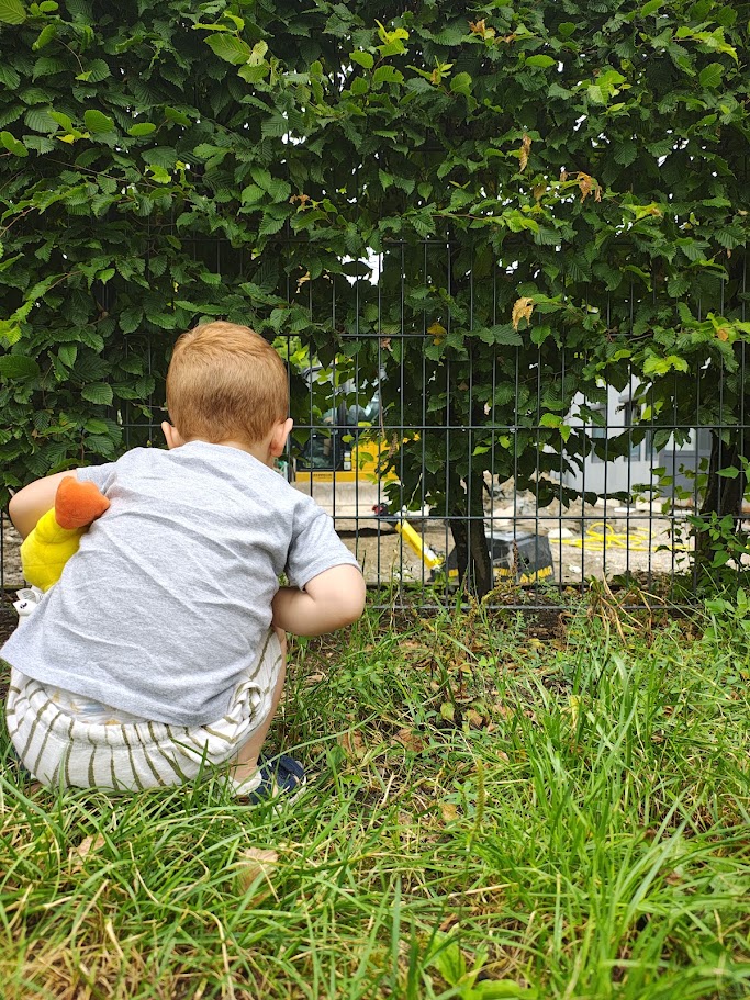 Un bambino accovacciato nell'erba sbircia un cantiere attraverso la siepe